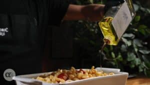 Person wearing a Chronic Guru apron drizzling cannabis-infused olive oil over a baked pasta dish with cherry tomatoes in a white casserole dish.