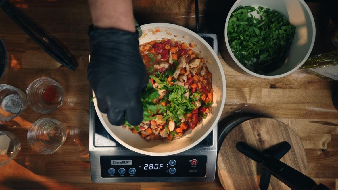 building the infused sweet potato chicken dinner, a gloved hand sprinkles in the chopped spinach