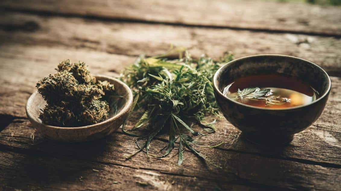 Bowl of cannabis buds, fresh leaves, and herbal tea on a rustic wooden table, symbolizing the connection between weed and digestion.